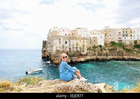 Touristen sitzen auf einer Klippe mit Blick auf das wunderschöne Polignano a Mare und das kristallklare Adria-Wasser in Italien. Stockfoto