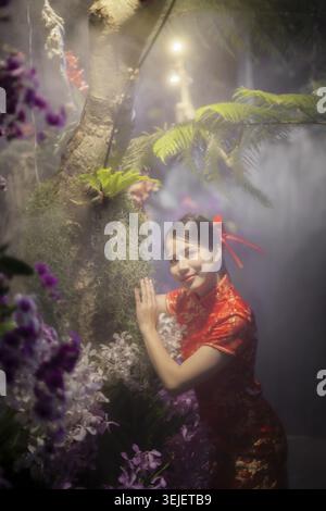 Schöne fröhliche asiatische Frau, die ein traditionelles rotes Cheongsam-Kleid trägt, posiert im nebeligen Fantasy-Garten mit Orchideenblume und Baum, der glücklich und glücklich aussieht Stockfoto