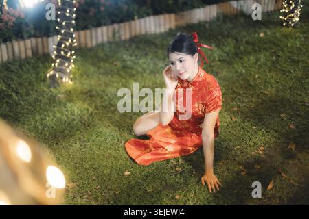 Elegante asiatische Frau mit rotem Cheongsam-Kleid, die nachts auf grünem Grasfeld im Freien sitzt und einen ruhigen und entspannten emotionalen Ausdruck zeigt Stockfoto