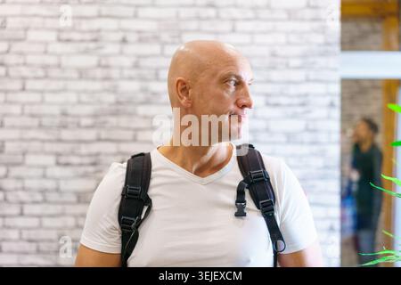 Glatze reife Männer Seitenprofil mit Rucksack vor weißer Ziegelwand in einem Café. Geeignet für maskuline Porträtmarken und urbane Innenausstattungen. Stockfoto