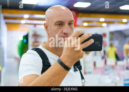 Glatze man macht Fotos auf dem Smartphone im Einkaufszentrum mit hellen Deckenleuchten. Geeignet für mobile Fotografie-Marken- und Einzelhandelstouren. Stockfoto