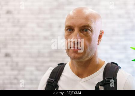Nahaufnahme des Glatzen Reifen Mannes mit Rucksack vor weißer Ziegelwand im Café. Geeignet für maskuline Markenwerbung und lässige Lifestyle-Inhalte. Stockfoto