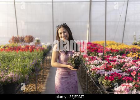 Glückliche junge asiatische Frau mit Topfblume in wunderschönem Gewächshaus. Shopping im Gartencenter voller farbenfroher Frühlingsblumen für schöne Gartenarbeit Stockfoto