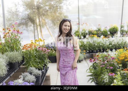Glückliche lächelnde asiatische Frau, die Blumen und Pflanzen im Sommergarten im Gewächshaus kauft. Wunderschöner Gast, der den Tag im Kinderzimmer genießt, sich fröhlich fühlt und sich wieder erholt Stockfoto