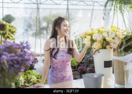 Glückliche junge Erwachsene asiatische Floristin lächelt in ihrem Blumenladen. Sie arrangiert fröhlich schöne Blumenstrauß in einem hellen und luftigen g Stockfoto