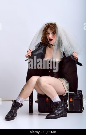 Junge schöne Frau überrascht mit verspieltem Braut-Look, der Leder und Eleganz in einem Studio kombiniert. Stockfoto