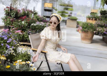 Stilvolle junge asiatische Frau, die sich ruhig und entspannt fühlt, sitzt auf dem Stuhl im Gewächshaus. Umgeben von bunten blühenden Blumen und grünen Pflanzen, genießt sie es Stockfoto