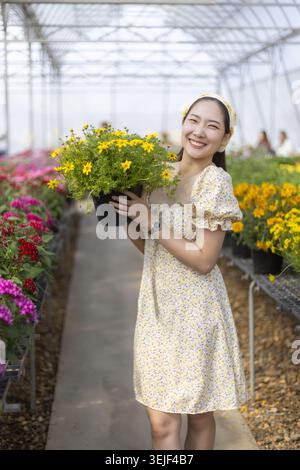 Glückliche junge asiatische Frau in hübschem Kleid, die einen Topf mit gelben Blumen im Gewächshaus hält. Diese fröhliche Gärtnerin lächelt beim Einkaufen von Colo Stockfoto