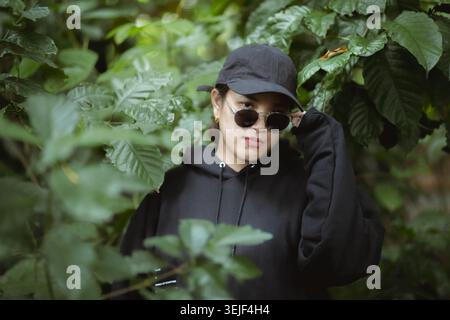Coole junge Erwachsene asiatische Person mit geheimnisvollem Ausdruck, die einen schwarzen Hoodie, eine Mütze und eine Sonnenbrille trägt. Porträt einer stilvollen Frau, die draußen im Stift steht Stockfoto