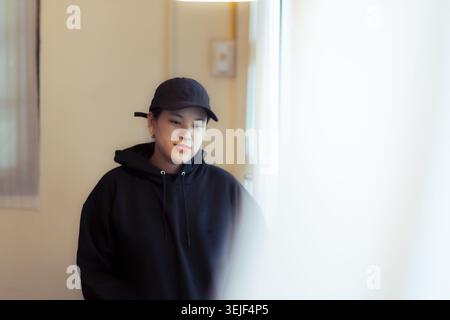 Junger asiatischer Mann mit schwarzem Kapuzenpullover und Mütze, der drinnen in der Nähe eines hellen Fensters steht und ruhige, besinnliche Emotionen zeigt, die sanft mit weichem li nach unten schauen Stockfoto
