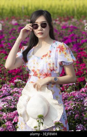 Selbstbewusste asiatische Frau, die in hellem Blumenfeld steht und schönes Blumenkleid und eine dunkle Sonnenbrille trägt, die einen Strohhut hält und sich ruhig und friedlich fühlt Stockfoto