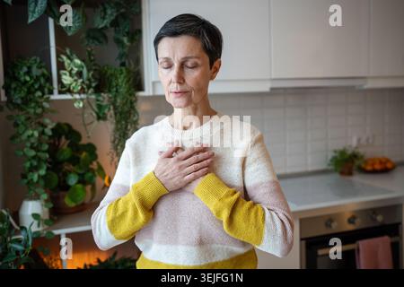 Fröhliche, ruhige, reife Frau, die mit tiefem Atmen meditiert, die Hände auf der Brust ruhen. Atmen zum Entspannen Stockfoto