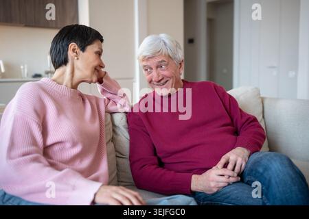 Glückliche Seniorenfamilie mit dem Eheleben. Ein lächelnder Ehemann erzählt Geschichten mit einer aufmerksamen Frau auf der Couch zu Hause Stockfoto