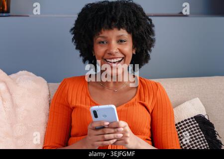Fröhliche schwarze Frau lacht, während sie zu Hause ein Smartphone auf einem Sofa benutzt Stockfoto