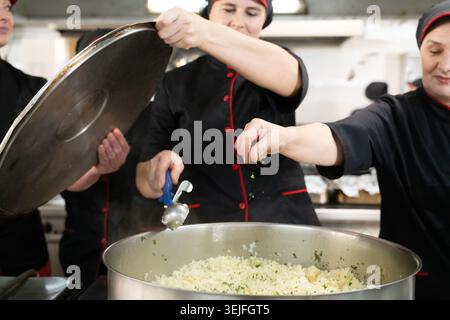 Gruppe von Köchen in schwarzen Uniformen, die Reis in einem großen Topf in einer kommerziellen Küche zubereiten. Weibliche Köche rühren und fügen Kräuter hinzu W Stockfoto