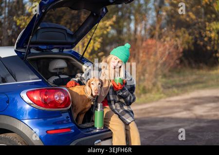 Frau trinkt Tee, während sie mit Hund im Kofferraum sitzt, Hund streichelt und die Natur im Wald genießt Stockfoto