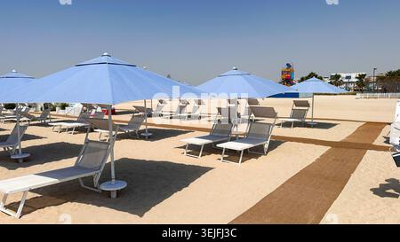 Reihe leerer Liegestühle und blauer Sonnenschirme, ordentlich am sonnigen Meer angeordnet, mit ruhigem Meer im Hintergrund. Stockfoto