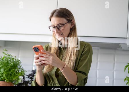 Glückliche Frau, die in entspannter Stimmung in der Küche auf dem Smartphone SMS schickt. Social-Media-Chat oder Online-Shopping Stockfoto