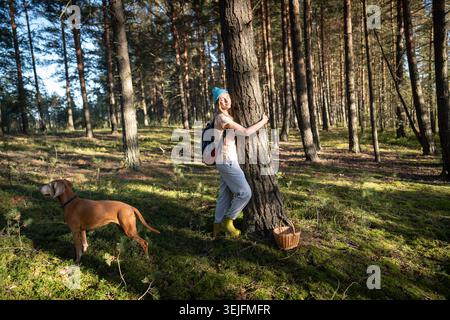 Entspannte Frau umarmt Baumstamm im Herbstwald mit geschlossenen Augen bei Sonnenuntergang, Spaziergang mit Hund im Wald Stockfoto