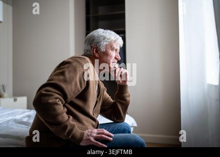 Seniorenmann allein zu Hause mit Hand über Mund, die in Angst und Apathie reflektiert. Demenzkrankheiten Stockfoto