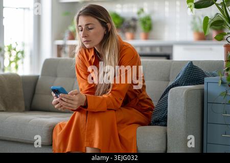 Frau mittleren Alters, die deutlich auf einen wichtigen Anruf wartet und auf den Smartphone-Bildschirm auf dem Sofa schaut Stockfoto