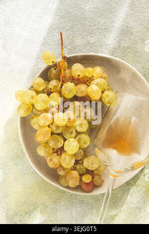 Ein Zweig weißer Trauben, auf einem grauen Teller, mit einem Glas Weißwein, Blick von oben, natürliches Licht, Lebensmittelkonzept, keine Leute, Stockfoto