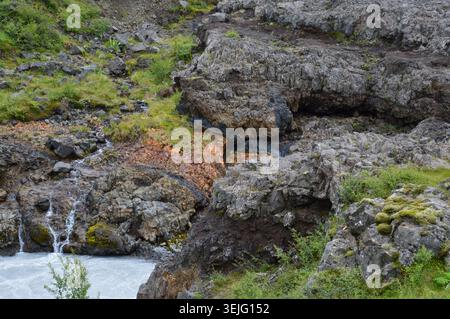 Ein detaillierter Blick auf geschichtete vulkanische Felsformationen und farbenfrohe Mineralsedimente auf einer zerklüfteten Klippe an einem rauschenden Fluss in Island. Stockfoto