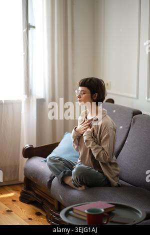 Junge Frau, die auf dem Sofa sitzt und Meditation praktiziert, Atemübungen für beruhigende Gedanken macht. Stockfoto