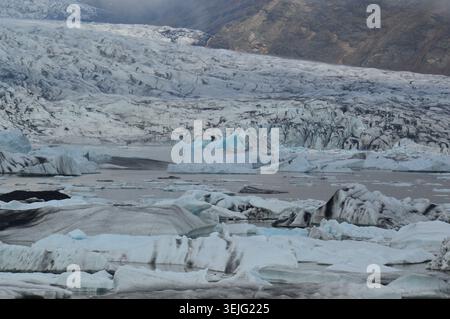 Großer blauer Eisberg mit ausgeprägten vulkanischen Ascheschichten, die im grauen Wasser einer isländischen Gletscherlagune schwimmen. Stockfoto