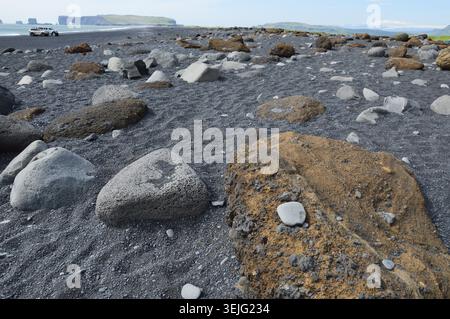 Verstreute vulkanische Felsbrocken und Felsen am schwarzen Sandstrand von Reynisfjara, Island, mit weit entfernten Landzungen. Stockfoto