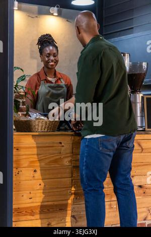 Afroamerikanischer Barista in grüner Schürze, der Gäste bedient, die an der Cafétheke mit Kartenleser zahlen Stockfoto