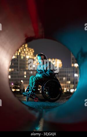 Nachdenklicher junger Mann im Rollstuhl in rgb-Neonlicht, der Zeit an der frischen Luft in der Nachtstadt verbringt. Stockfoto