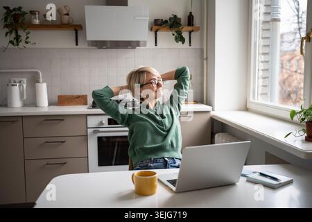 Fröhliche Geschäftsfrau, die sich im Stuhl zurücklehnt, schaut ins Fenster und fühlt sich mit der Arbeit zufrieden. Stockfoto
