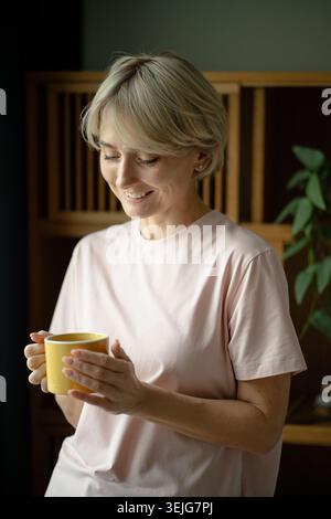 Lächelnde Frau mittleren Alters im Pyjama beginnt früh am Morgen mit einer Tasse aromatischem Kaffee und Positivitäten Stockfoto
