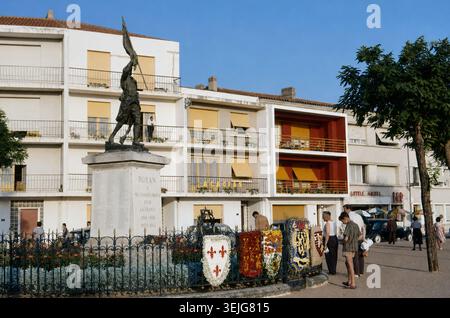 Frankreich der 1960er Jahre in Farbe - Kriegsdenkmal in Royan, Charente-Maritime, fotografiert 1963. Bronzestatue eines Soldaten, der die französische Flagge auf einem Steinpodest hebt, mit der Inschrift „Royan à ses enfants morts pour la France 1914–1918, 1939–1945“, die an die Toten des örtlichen Krieges erinnert. Die umliegenden Eisengeländer sind mit Blumenschildern geschmückt, mit Apartmentgebäuden aus der Mitte des Jahrhunderts, Hotelschildern und Fußgängern in Sommerkleidung, die das französische Küstenleben der Nachkriegszeit widerspiegelt. Stockfoto