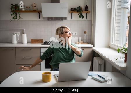 Verschlafene Geschäftsfrau mittleren Alters, gähnt und dehnt sich, sitzt am Tisch und arbeitet von zu Hause aus am Laptop. Stockfoto