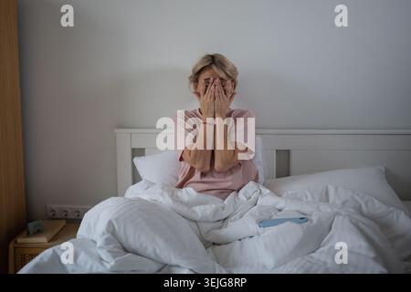 Verschlafene Frau mittleren Alters, die nach einer unruhigen Nacht im Bett sitzt und mit Kopfschmerzen und Müdigkeit zu kämpfen hat. Stockfoto