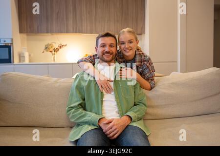 Fröhliches, nettes Paar von Mann und Frau, die sich umarmen und lächeln und sich vor der Kamera im gemütlichen Wohnzimmer posieren. Stockfoto