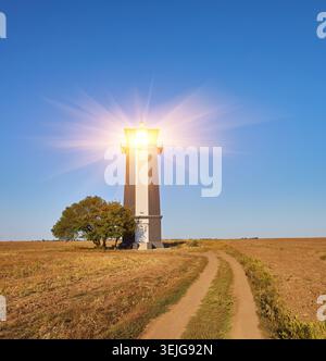 Ein hoher, gestreifter Leuchtturm (rote, weiße und möglicherweise schwarze/dunkle Bänder), der auf einem großen, offenen, trockenen Feld unter einem klaren blauen Himmel steht. Ein einzelner Baum Stockfoto