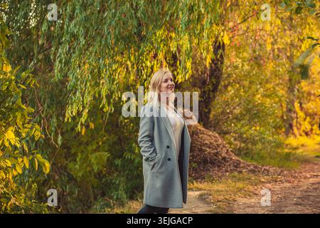 Junge blonde Frau im stilvollen grauen Mantel, die im goldenen Herbst auf dem Naturparkweg spaziert. Warmes Sonnenlicht beleuchtet sattes grüngelbes Laub Stockfoto