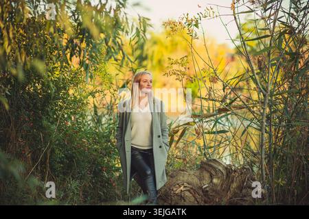 Blonde Frau in stilvollem grauen Mantel schwarze Lederhose steht neben ruhigem Wasser. Umgeben von leuchtend grünen, gelben Herbstblättern blickt sie jedoch Stockfoto
