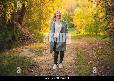 Die fröhliche blonde Frau in grauem Mantel und schwarzer Hose spaziert durch den sonnendurchfluteten Herbstpark. Warmes goldenes Licht leuchtet leuchtend gelb und grün auf Stockfoto