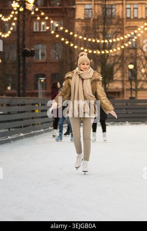 Junge Frau im Pelzmantel, beigefarbener Hut mit Pompon, Schal Schlittschuhlaufen auf Eislaufbahn, im Winter draußen Stockfoto