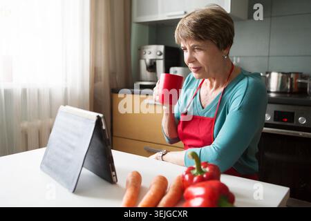 Elegante ältere Frau, die Tablette in der Küche benutzt Stockfoto