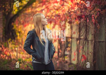 Eine nachdenkliche junge Frau mit blonden Haaren steht im Profil. Sie trägt lässige Herbstmode, Hände in Taschen ihrer schwarzen Lederjacke Stockfoto