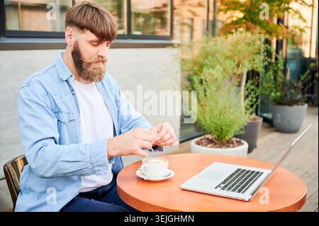 Bärtiger Mann rührt Kaffee neben Laptop am Tisch auf der Außenterrasse und genießt eine entspannte Fernarbeitspause zwischen Pflanzen und moderner Architektur Stockfoto