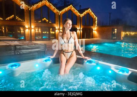 Weibliche Entspannung im beleuchteten Whirlpool vor dem modernen Holzhaus, festliche Lichter und Winteratmosphäre, Wellness-Lifestyle und luxuriöses Rückzugskonzept Stockfoto