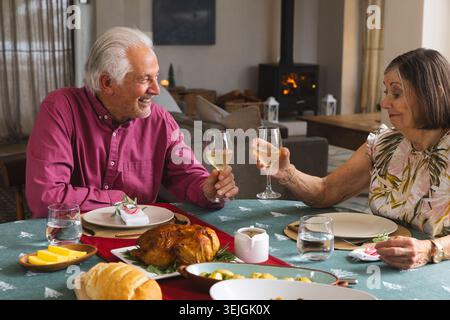 Seniorenpaar in burgunderrotem Hemd und Bluse, die Weingläser am Esstisch zu Hause mit Geflügel hochziehen. Intimität, Feier, Wärme Stockfoto