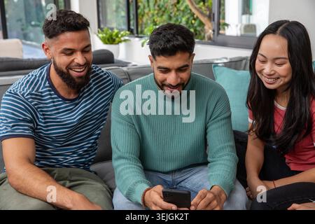 Verschiedene Freunde sitzen auf grauem Sofa im Wohnzimmer, Mittelmann in grünem Pullover mit Smartphone. Sofa, Gruppe, Tageslicht, Innenraum, Grün, Anlage Stockfoto