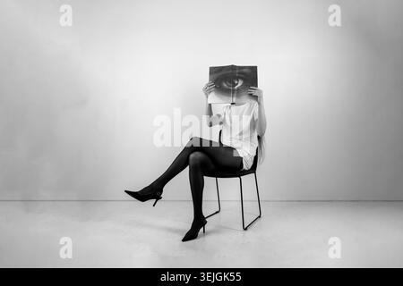 Frau, die auf einem Stuhl sitzt und ein offenes Buch mit einem Augenabdruck über dem Gesicht hält Stockfoto